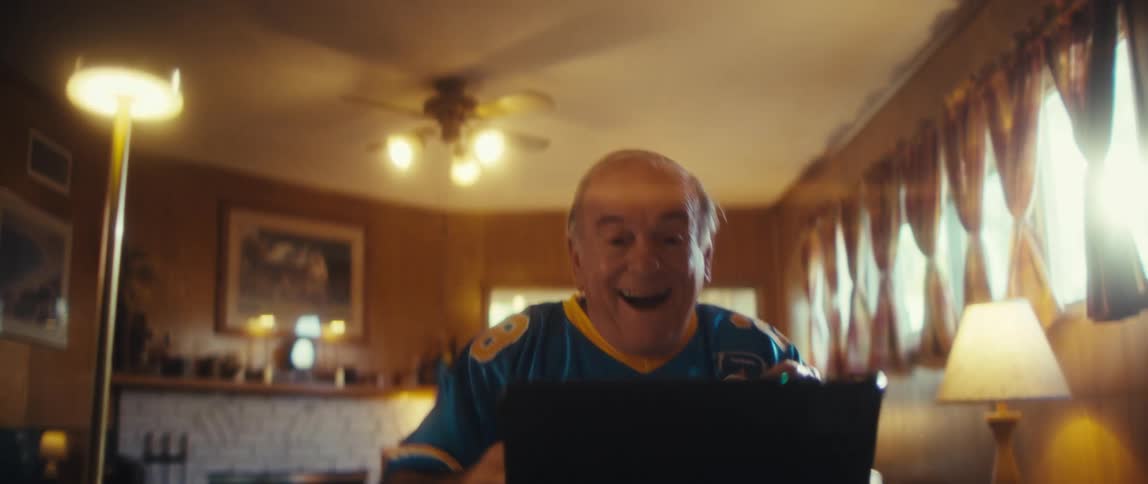 An elderly man wearing a team jersey sits in front of a tablet cheering happily