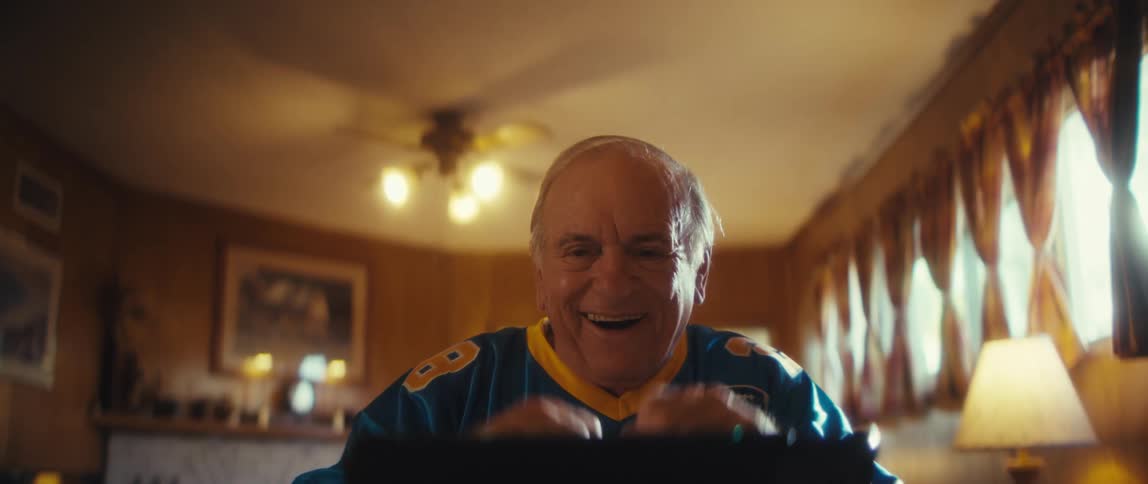 An elderly man wearing a sports jersey is watching a tablet in front of him while waving his arms and smiling in a sunlit room