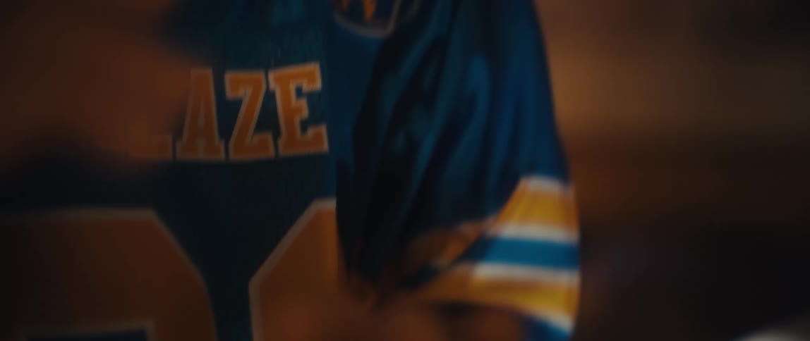 A male wearing a blue and yellow garment claps his hands together and smiles widely while standing indoors with bright backlighting