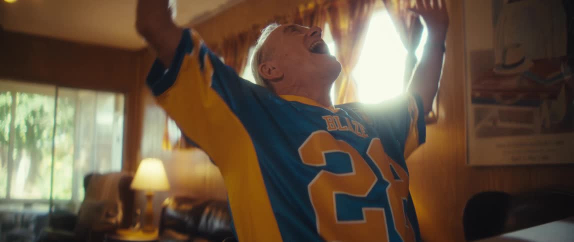 An ecstatic elderly man in a sports jersey joyfully celebrates a victory raising his arms and laughing in a sunlit living room