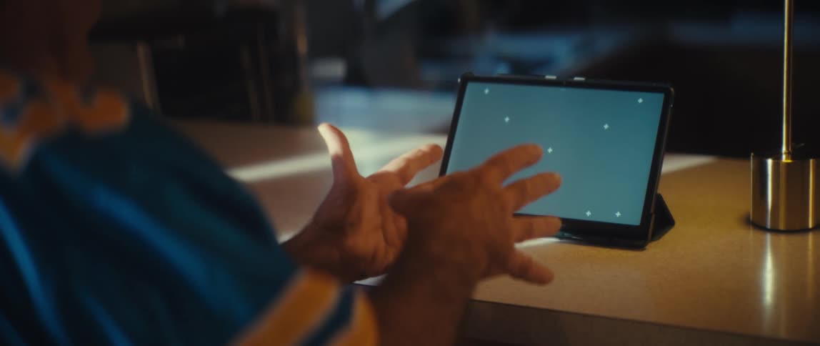 From behind a closeup shows an older man wearing a blue and yellow jersey with legend printed on the back he is sitting at a desk enthusiastically clapping and cheering while looking at a tablet with a green screen chromakey di