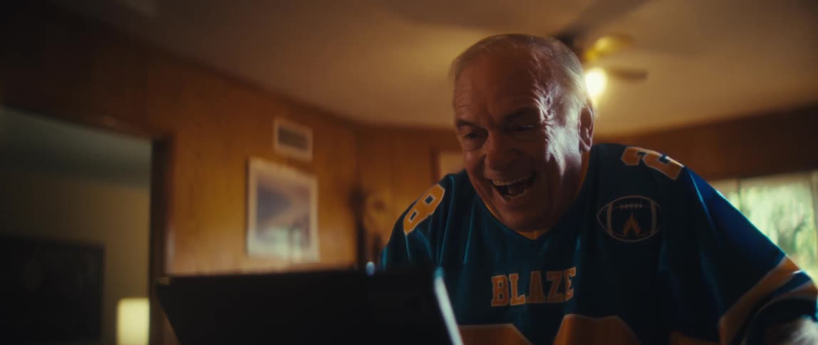 An elderly man wearing a jersey watches a game in his living room suddenly bursting with excitement