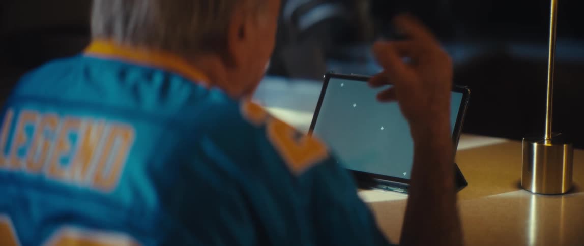 A close view video from behind shows an older man with gray hair wearing a blue and yellow sports jersey that says legend as he gestures while looking intently at a tablet device with a chroma key screen placed on a dark wooden desk