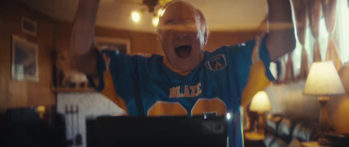 A happy older man with light hair wearing a blue and yellow sports jersey reacts with excitement and raises his hands while looking at a black laptop or tablet in a warmly lit woodpaneled living room with a picture frame and mantel in the background