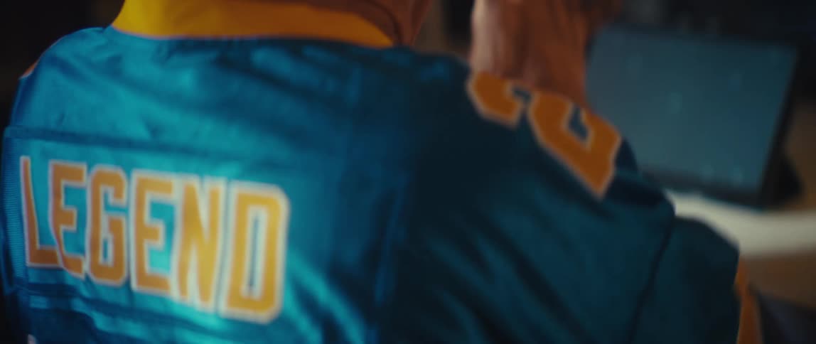 A tight shot focuses on the back of a turquoise and yellow sports jersey that has the word legend printed across the shoulders the wearer of the jersey turns to sit down at a table with a tablet propped up on it its screen showing a blan