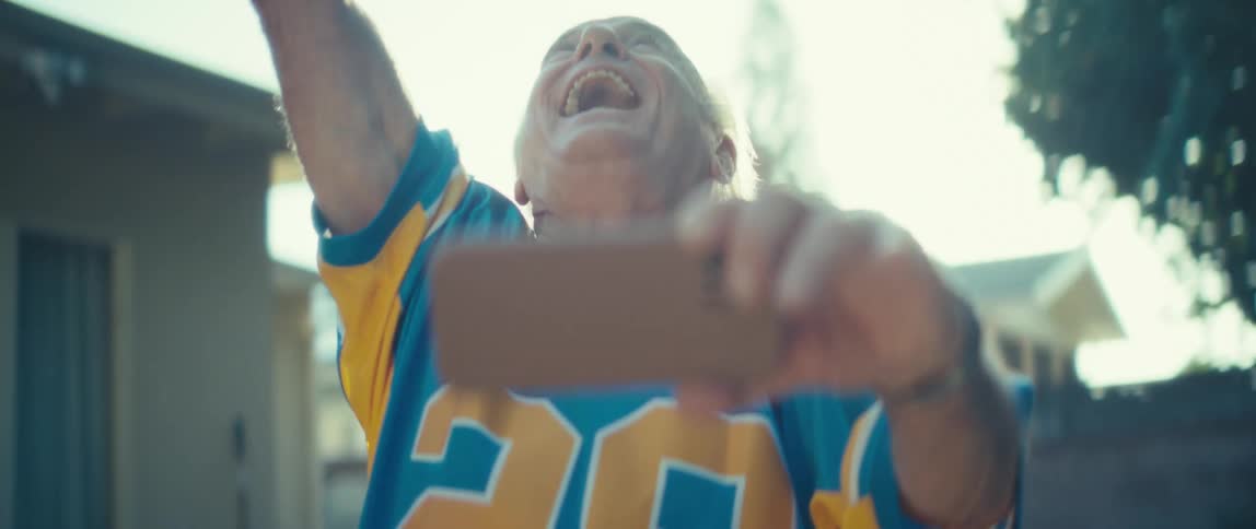 An elderly man stands outdoors at sunset holding his phone and suddenly bursting with excitement while wearing a jersey