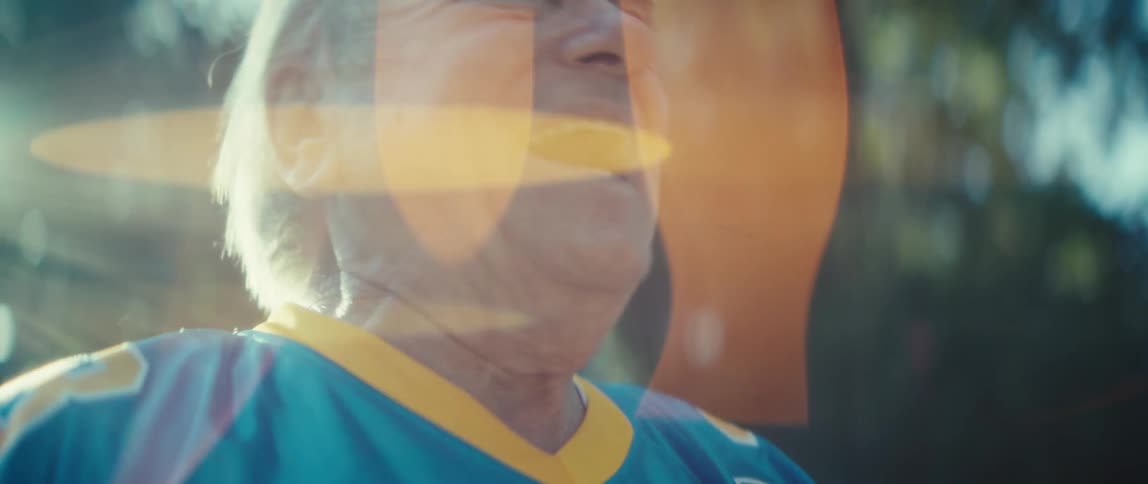 A sundrenched closeup captures the older mans face as he laughs and cheers with an ecstatic openmouthed smile he is looking up and raising his arms in excitement with a bright lens flare washing over the frame