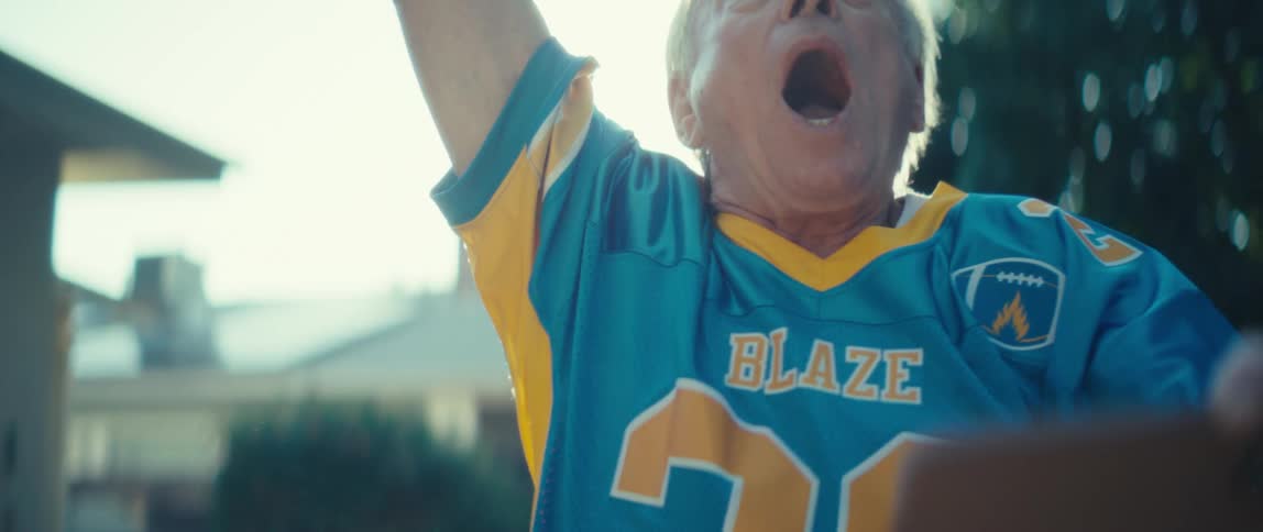A brightly lit closeup shows an older man wearing a blue and yellow sports jersey with blaze on the chest he looks down reacts with a surprised openmouthed expression and then raises his arms and shouts in excitement