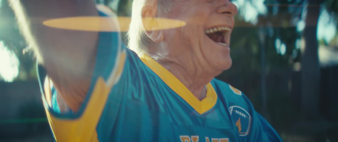 An older male in a sports jersey holds a mobile phone shouts with his mouth open and raises both arms in a triumphant gesture while standing outdoors