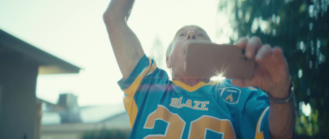 An elderly man in a blue blaze 20 sports jersey raises a triumphant arm beaming with joy as he looks at a phone he is holding