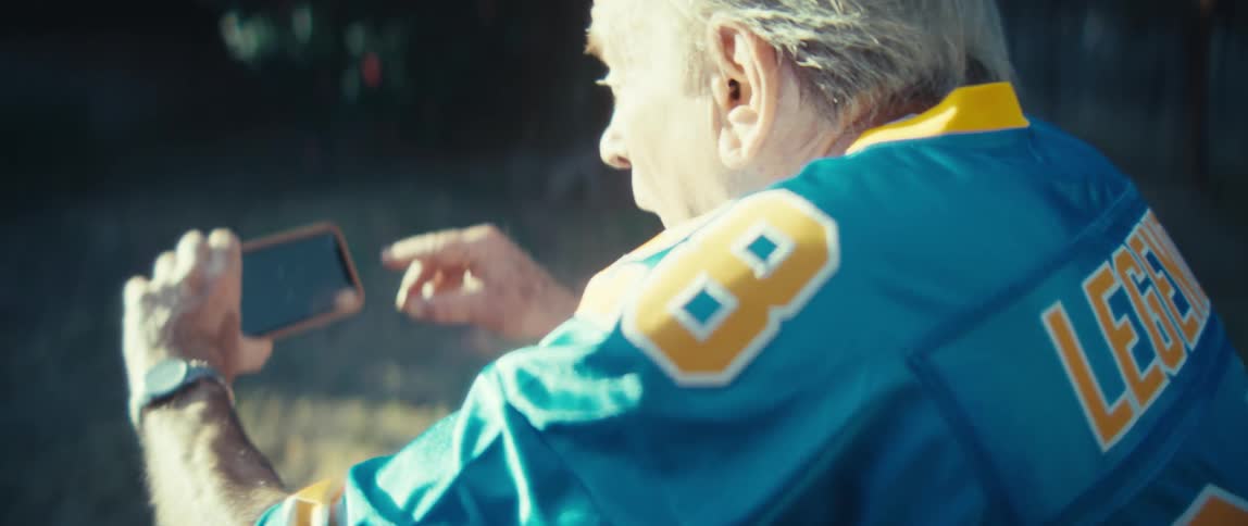 An elderly man wearing a sports jersey cheers in a backyard while watching something on a tablet