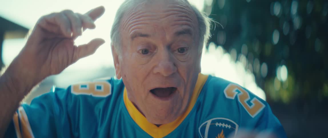 A close view video shows an older man wearing a blue and yellow sports jersey holding a smartphone horizontally with his mouth open in surprise before he smiles in victory or excitement in the bright sunny outdoor setting