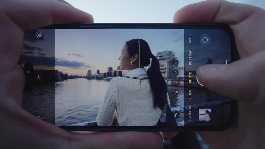 Hands hold a smartphone camera focusing on a woman by a waterfront skyline female appearing calm and contemplative