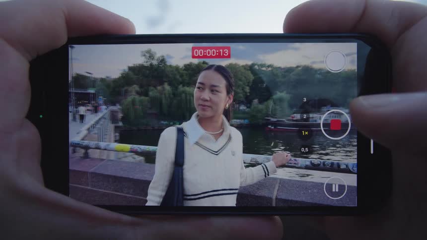 Two human hands hold a smartphone displaying its video camera application which records an asian female human being standing by a water channel who turns toward the lens and smiles