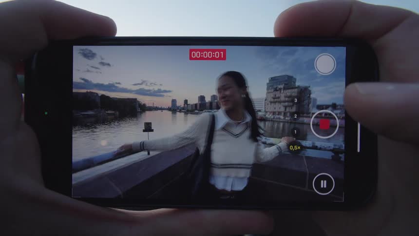 A persons hands hold a smartphone horizontally showing a close view video recording of a smiling young woman in a white sweater vest standing on a bridge railing with a river and city buildings visible in the background against a dusk sky