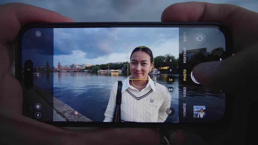 Two human hands hold a smartphone displaying its camera application which focuses on an asian female human being standing by a waterfront and turning to smile at the lens in a city setting