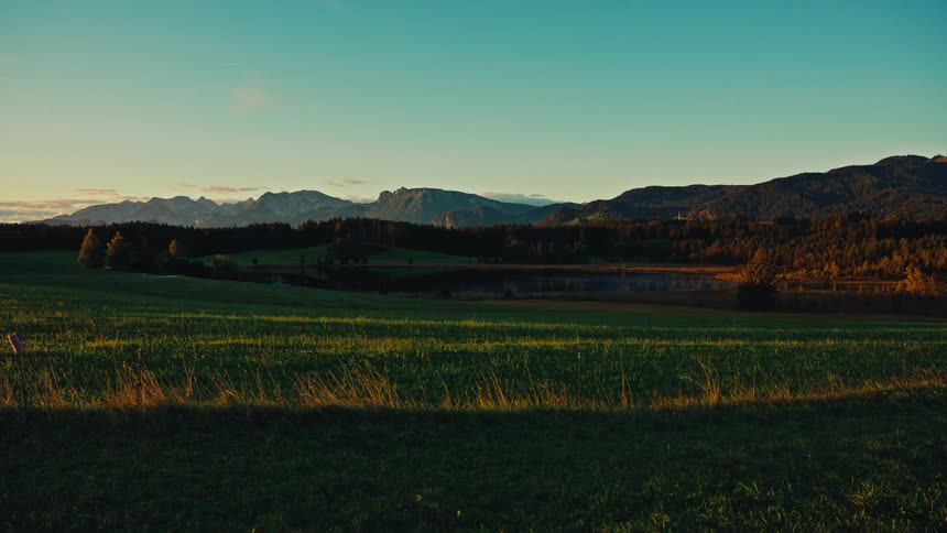 An empty sunlit grassy field stretches toward distant mountains