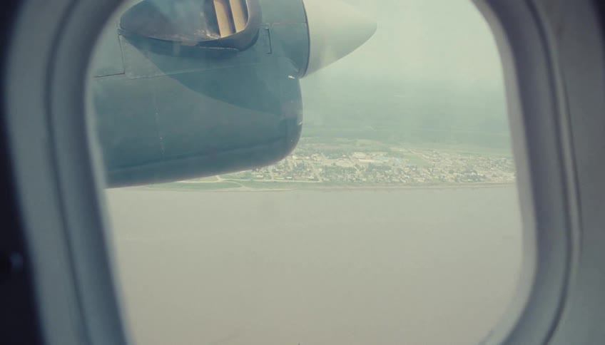 A view through an oval airplane window shows the propeller engine housing as the aircraft descends over a body of water and reveals a scattered settlement of buildings on the land below