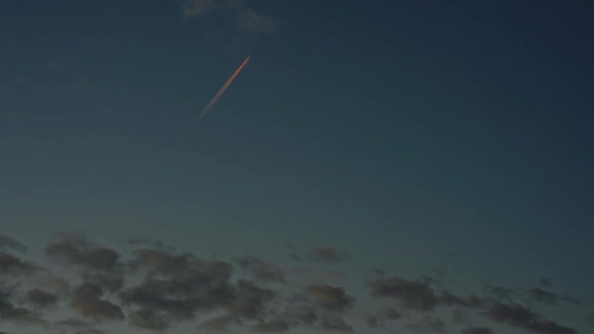 Fading sunlight tints the contrail created by a jet flying above dark clouds