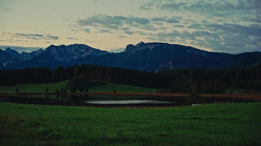 A wide shot shows a field of grass extending to a small body of water bordered by a forest of dark trees with a large high mountain range visible beyond the tree line beneath a cloudy sky