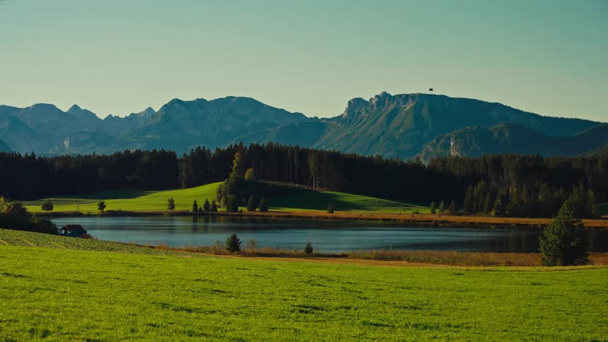 A stunning wideangle shot captures a serene mountain lake nestled in a valley reflecting the blue sky and surrounding dark green forest the foreground features a vibrant sunny green meadow sharply contrasting with the craggy bluegre