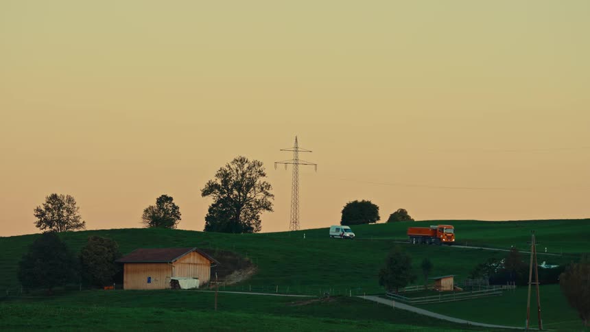 A static long shot captures a rural hilly landscape bathed in the golden glow of sunset or sunrise a prominent power line tower stands on one of the bright green rolling hills while an orange dump truck and a white van drive slowly on