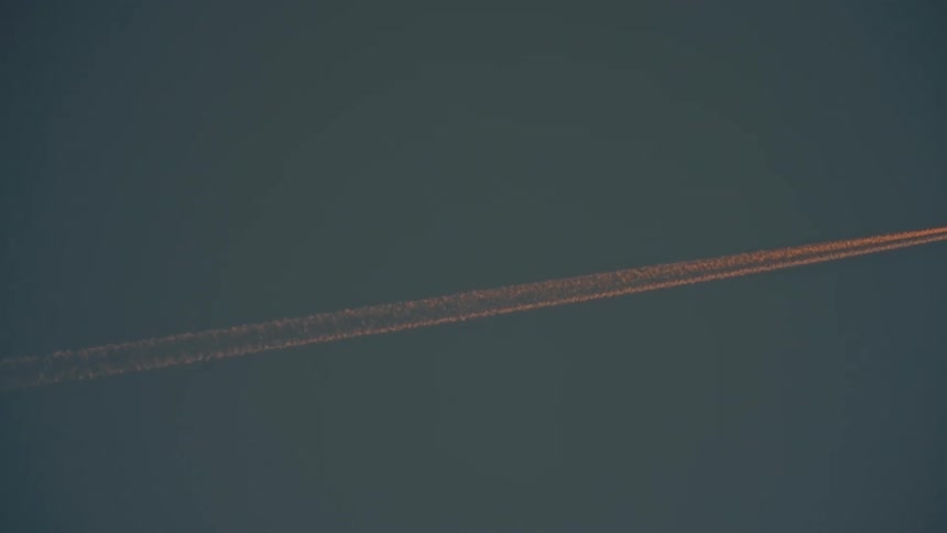 An airplane flies across the sky at midday leaving orange trails behind it