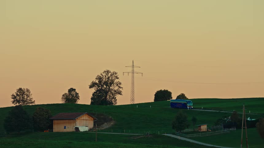A timelapse video shows a blue tour bus and a white car quickly driving along a winding road through rolling green hills with a few farm buildings and a large metal electricity pylon silhouetted against a pale orange sunset sky