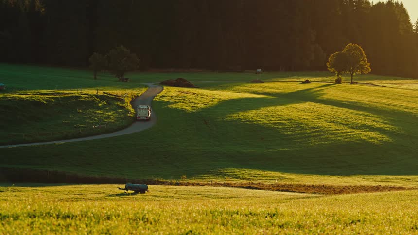A small white delivery truck drives slowly up a winding dirt road through rolling green hills bathed in the warm deep orange light of a sunset toward a dark evergreen forest silhouetted against the bright sky