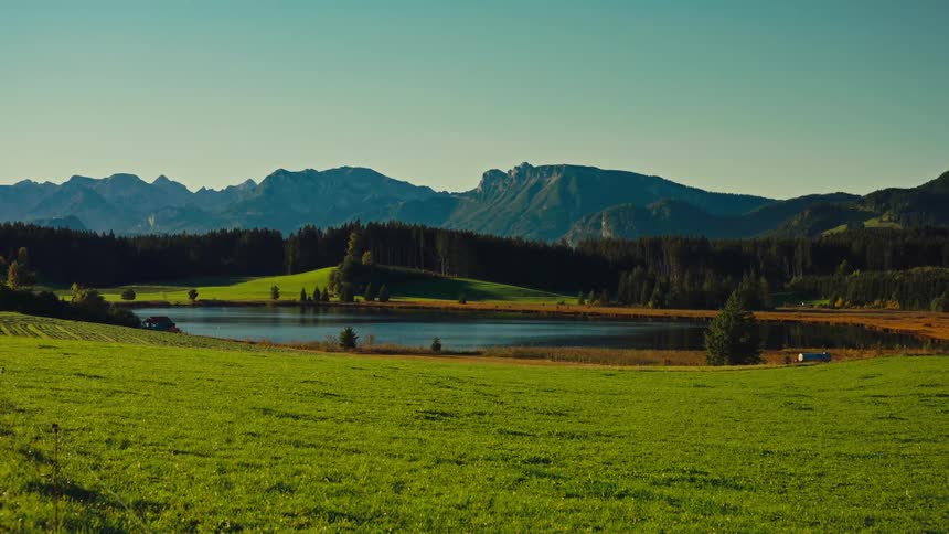 A stunning long shot captures a bright green sunny meadow leading down to a dark peaceful mountain lake beyond the lake a dense evergreen forest covers a rolling hillside which is backed by a majestic rocky bluegrey mountain range