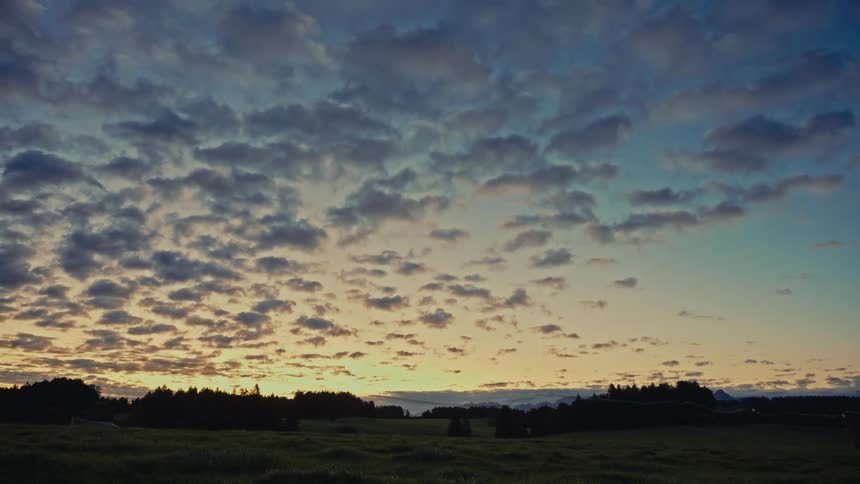 A wide shot of an outdoor environment shows a horizon of dark trees and fields beneath a sky filled with patchy clouds that slowly change color from blue to orange and yellow