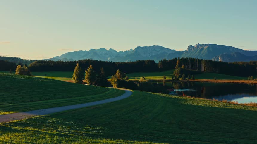 An aerial perspective of a grassy hill with a curving paved path descends toward a still lake which is bordered by a dense forest and backed by a tall mountain range under a clear sky with golden light