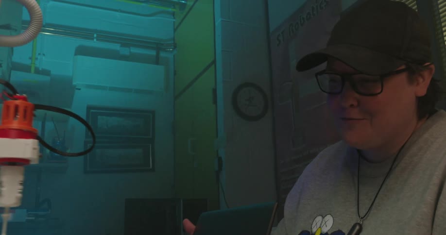 A dark midshot captures a person wearing a baseball cap and glasses working at a desk next to a robotic arm the low light is tinted blue by the environment highlighting the person and the brightlylit robotic arm