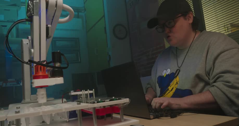 A young man wearing glasses sits at a desk and types on a portable computer while monitoring a small white robotic arm that is operating a scientific device in an indoor workspace