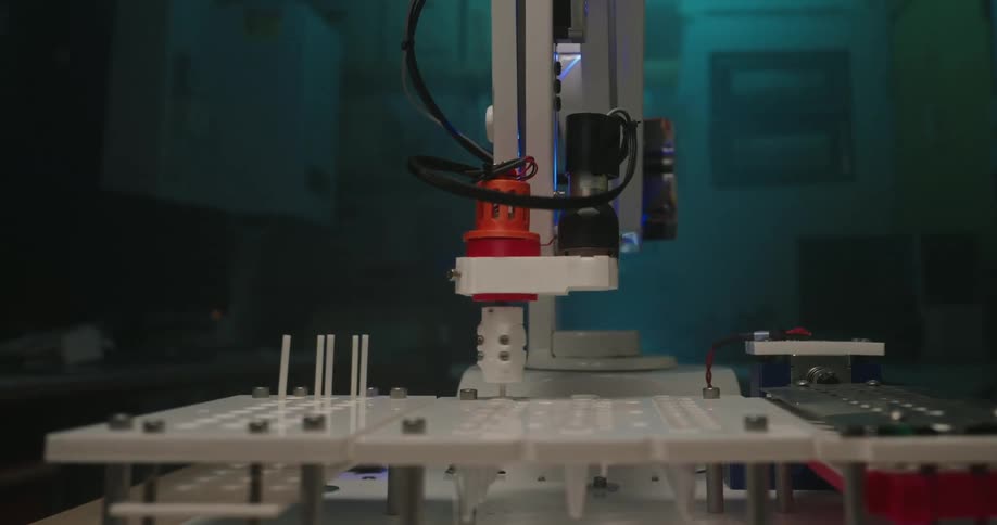 A small white robotic arm descends and rotates to perform an operation over a white testing platform inside a dimly lit room with blue illumination
