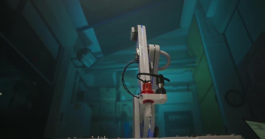 A white and gray robotic arm with a red tip and blue led lights moves in a slow precise motion in a dark futuristic laboratory or industrial setting with a bluegreen ambient glow