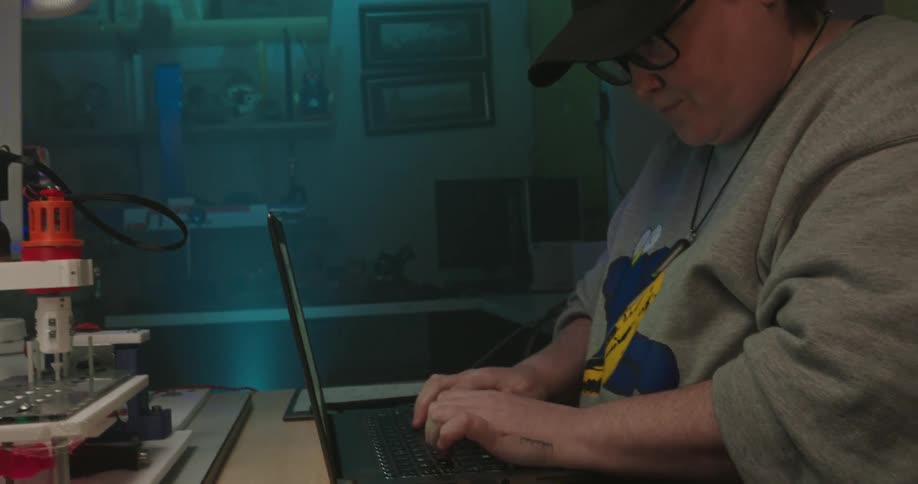 Person wearing a cap and glasses types on a laptop beside electronic equipment on a workbench