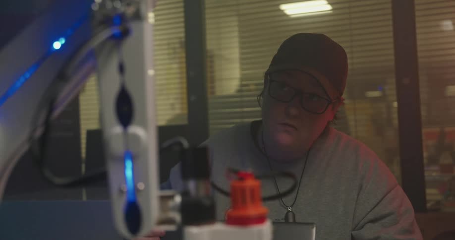 An overhead medium shot focuses on a person wearing a black beanie and glasses sitting at a desk in the foreground a robotic arm with blue glowing accents is partially visible suggesting a technological or industrial setting
