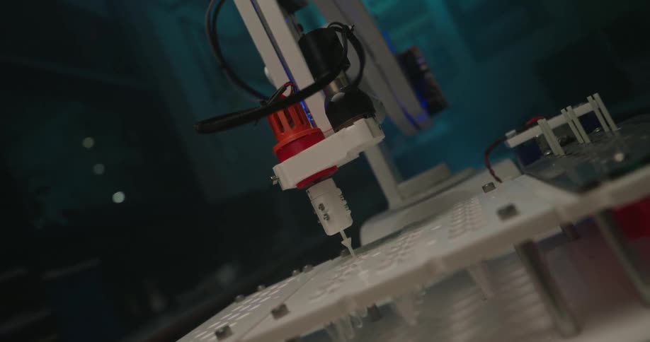 A medium slightly lowangle shot focuses on a robotic arm with a red and white dispenser attachment working on a white mechanism on a desk the overall scene is dark and tinted with a cool blue light