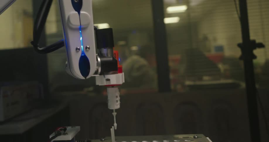 A midcloseup shot shows the white and blue arm of a robotic device with a syringe or dispensing nozzle on the end the robot is moving slowly against a dark industriallooking background with muted long fluorescent lights visible in the ceilin