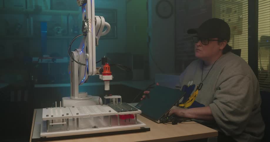 In a dimly lit workshop a person at a laptop operates a sophisticated robotic arm mounted on a desk