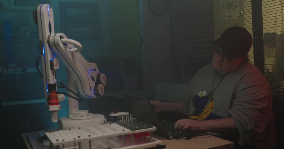 The video shows a medium shot of a person sitting in a dimly lit space likely a laboratory or workshop operating a laptop with a complex white robotic arm next to themthe person is wearing glasses a black baseball cap and a grey sweatshirt with