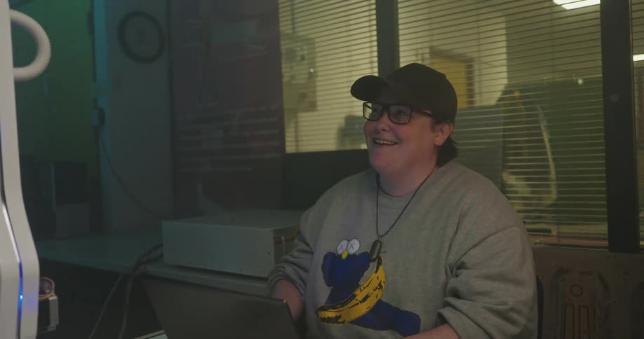 A young man wearing a cap and glasses sits at a desk smiles while typing on a portable computer and occasionally looks toward a small robotic device in an indoor room