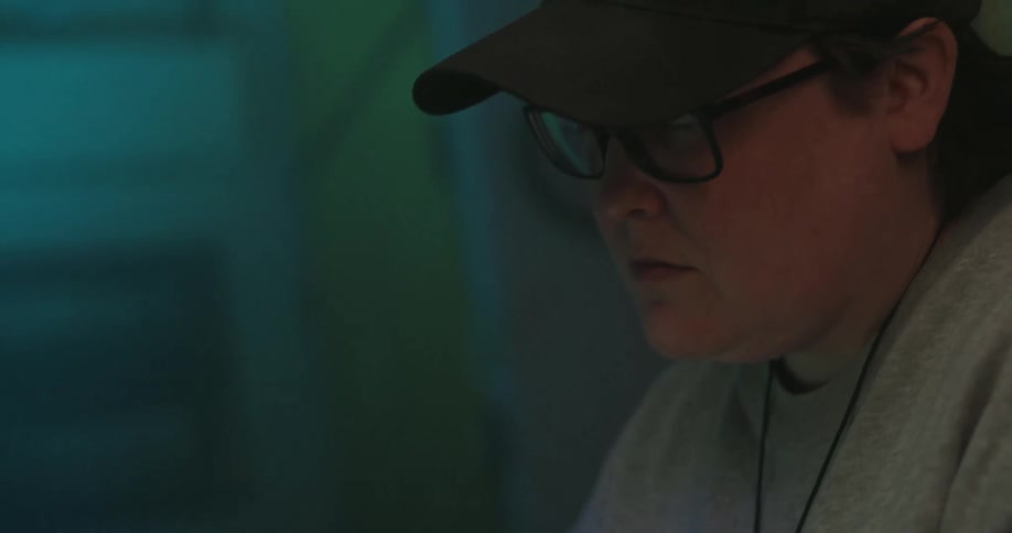 A young woman in glasses and a hat sits behind a desk typing on her laptop