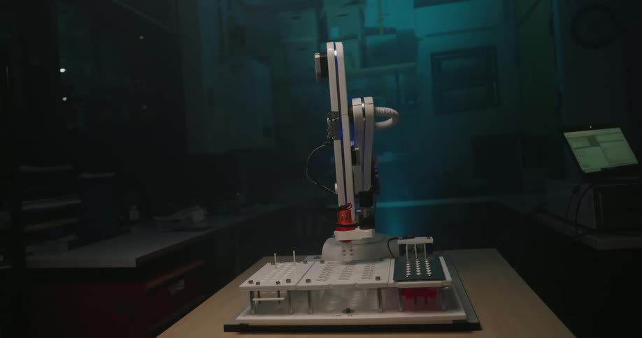 The video features a small white robotic arm likely a lab automation or educational model mounted on a table in a dimly lit possibly industrial or makeshift laboratory setting the background is shrouded in bluegreen atmospheric lighting