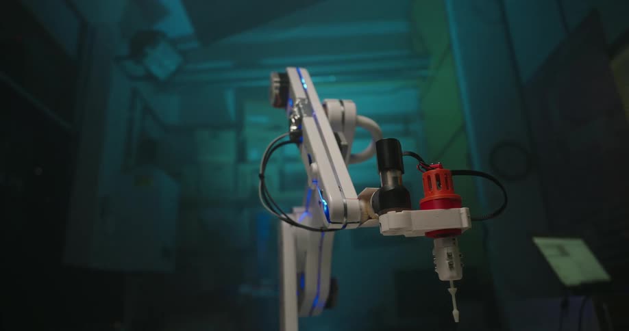 Robotic arm with red gripper operates over surface with circular cavities in lab setting with monitors in the background