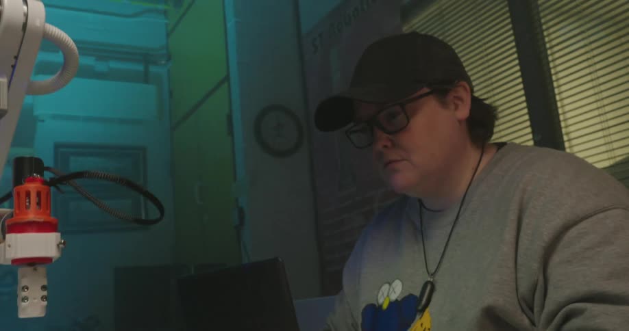 A young man wearing a baseball cap and glasses sits at a desk and operates a laptop while looking between the screen and a small white robotic machine to the side