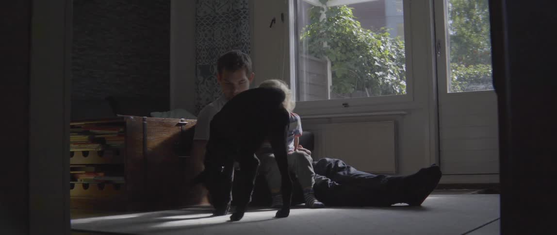 A father and toddler sit on the floor playing with a large black dog.