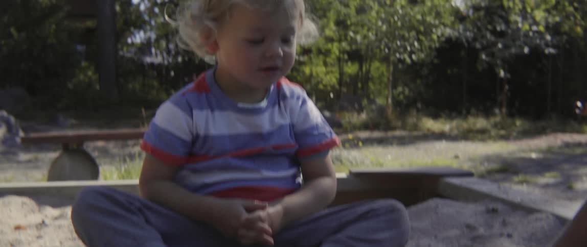 Young toddler boy sitting in a sandbox and looking up at his mother curiously.