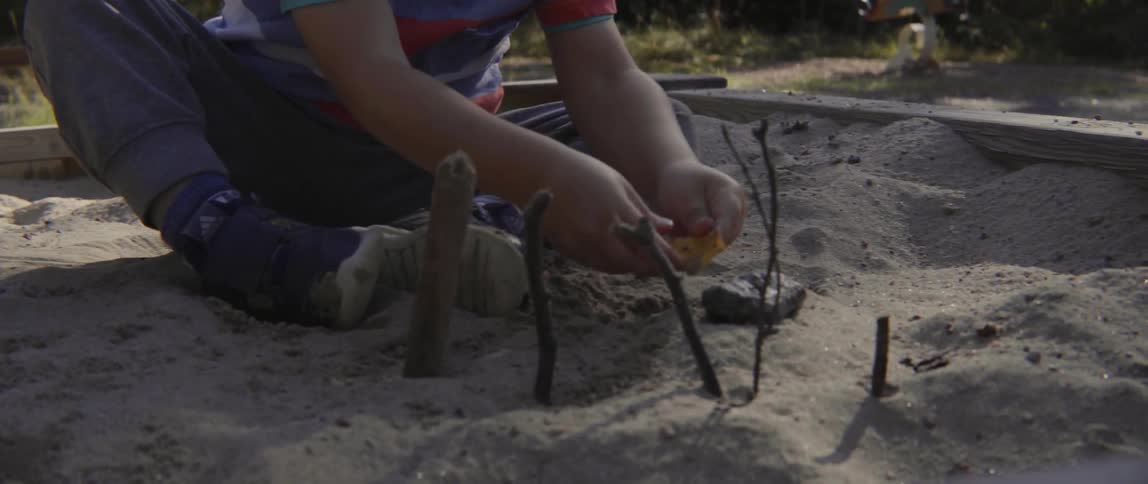 Young toddler boy focused on sensory play while sitting in a wooden sand pit.
